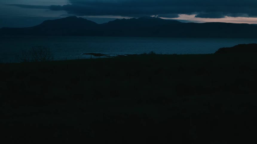 A somber, wide shot of a dark lake and mountains at dusk