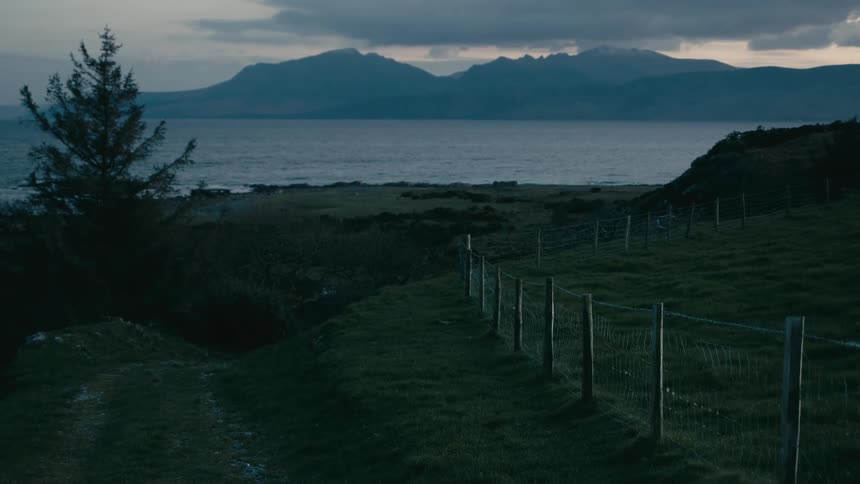 A tranquil and moody view of a rugged coastline at dusk, with a grassy hill and fence line overlooking the sea and distant mountains