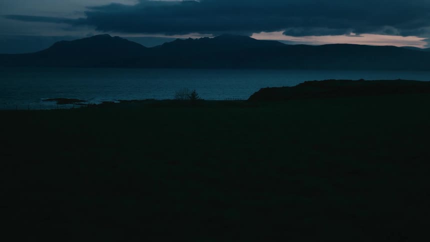 A dark and moody view of a silhouetted coastline with mountains across the water under a cloudy twilight sky