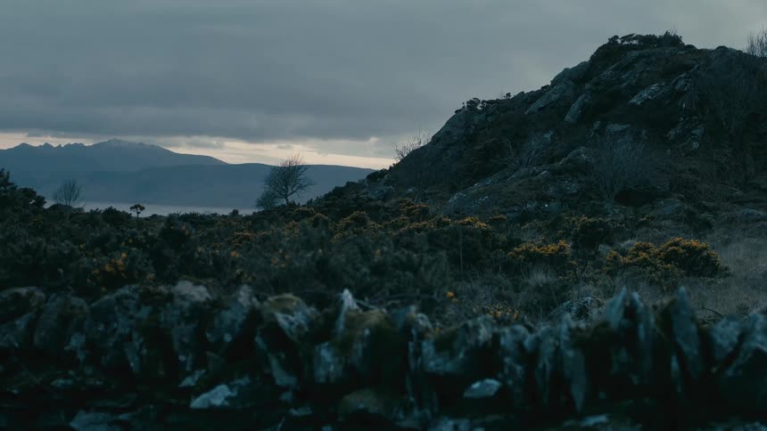 A gloomy, overcast landscape view over a mossy stone wall of a rocky hillside with snowy mountains in the distance