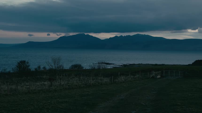 A moody and atmospheric view of a calm sea and distant mountains under a dark, cloudy sky at dusk