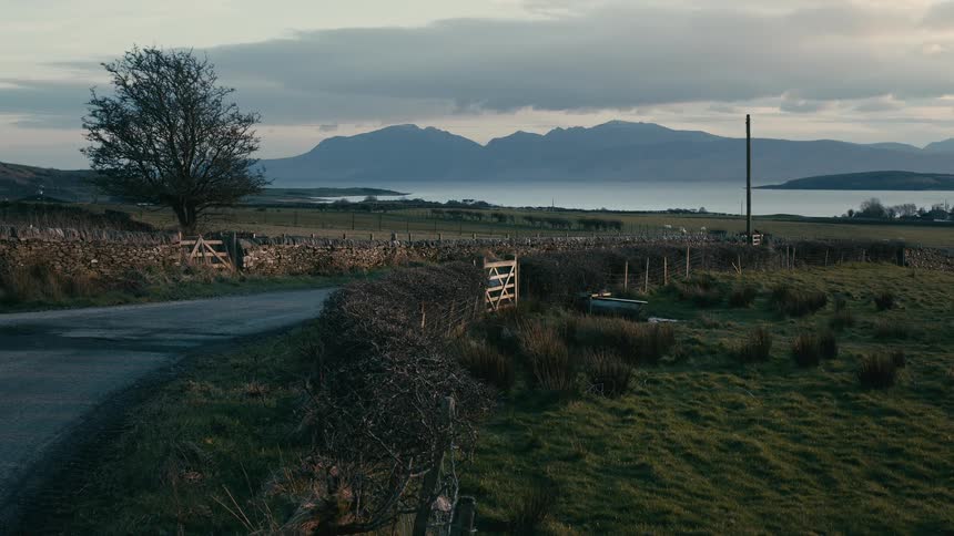 A peaceful rural landscape featuring a country road, stone walls, and a scenic view of mountains across the water at dusk