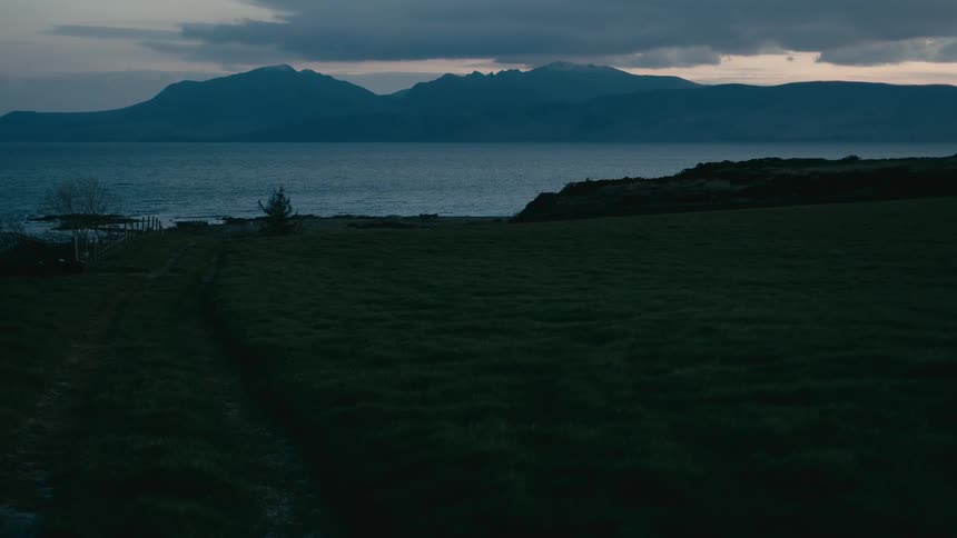 A tranquil twilight view across a grassy field to a still loch and majestic mountains under a cloudy sky