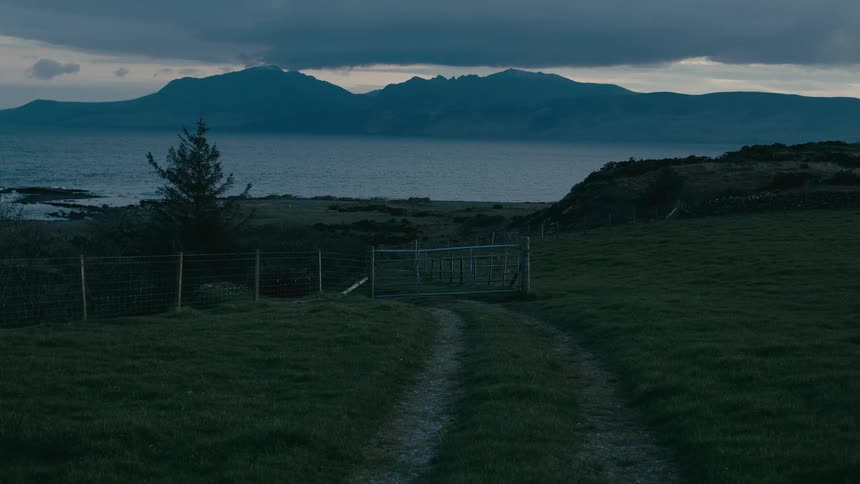 A dirt path leads through a green pasture towards a gate by the water, with mountains visible in the distance at dusk