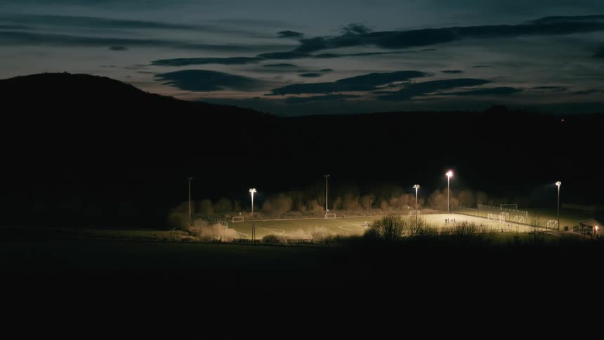 A wide, static shot of a brightly lit soccer field at night where a team is practicing in a dark, rural valley