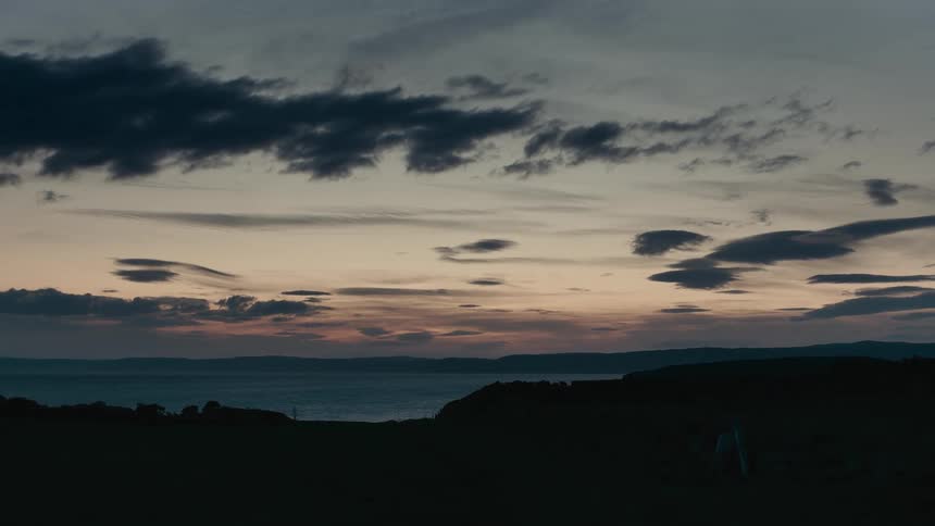 A tranquil view of the sea from a silhouetted coastline under a dramatic cloudy sky at dusk