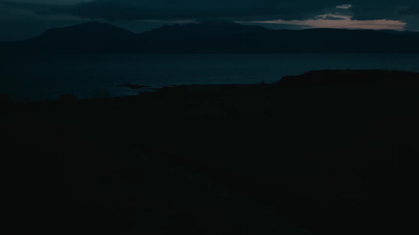 A moody, dark shot of a quiet shoreline with distant mountains silhouetted against the twilight sky