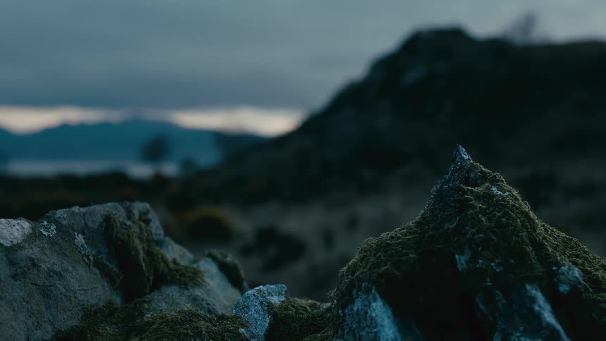 An atmospheric close-up of a mossy stone wall overlooks a vast, blurry mountain landscape at dusk