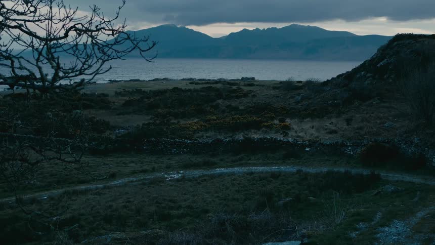 An atmospheric view of a rugged, remote coastline with mountains across the water under a moody, overcast sky