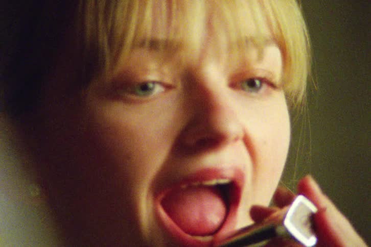A grainy, film-style close-up of a stylish young blonde woman applying red lipstick