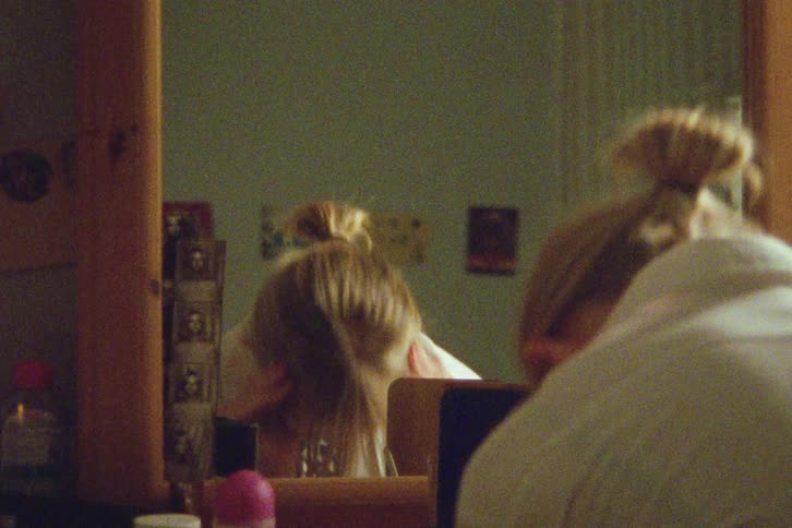 Over-the-shoulder shot of a young woman with earbuds looking in the mirror and styling her hair in a vintage-style room
