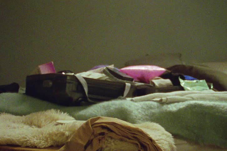 A woman in vintage attire chaotically packs her suitcase on a bed, dancing and moving around the room in a grainy, filmic style