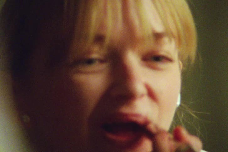 A grainy, vintage-style close-up shot of a blonde woman with bangs applying lipstick in a dimly lit room