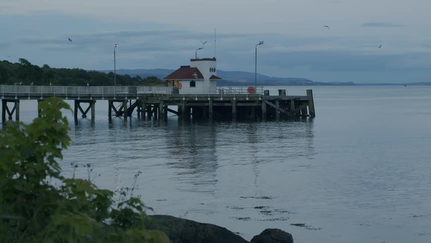 A tranquil pier with a white building extends over calm water as seagulls fly overhead on a grey, overcast evening