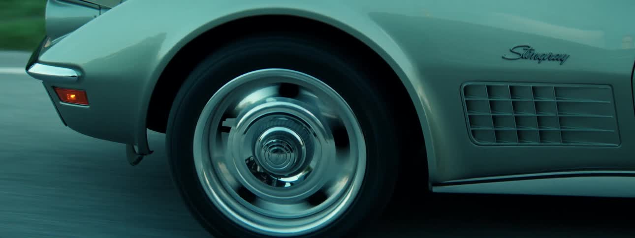 A moody close-up shot captures the spinning chrome rear wheel of a vintage silver Corvette as it speeds down the road