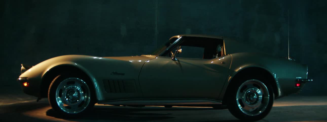 A classic silver sports car is parked in a dark, cinematic setting, with a single light creating dramatic highlights