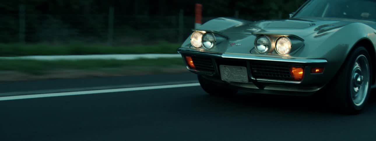 A cinematic close-up of a vintage silver Corvette with its pop-up headlights on as it drives down a highway at dusk