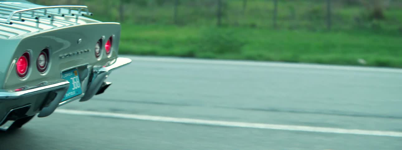 A stylish tracking shot follows the rear of a classic silver-green Chevrolet Corvette as it drives along a highway