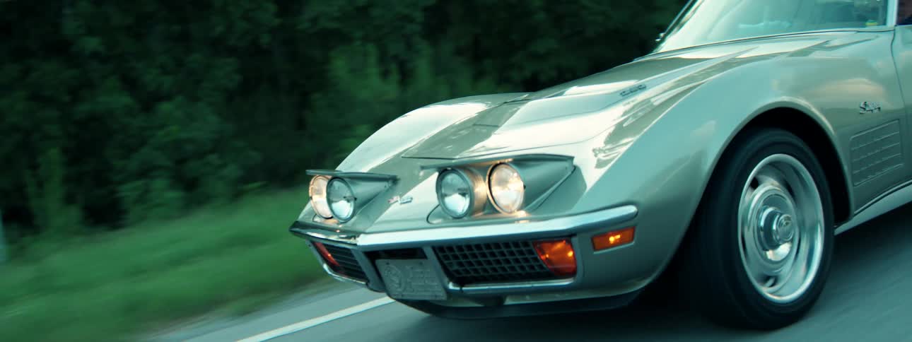 A low-angle close-up shot follows a classic silver-green sports car with its pop-up headlights on as it drives along a highway