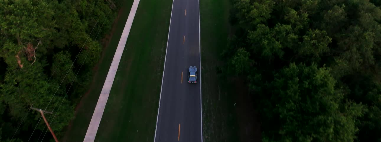 An aerial top-down view tracks a classic DeLorean time machine car driving along a quiet, tree-lined country road