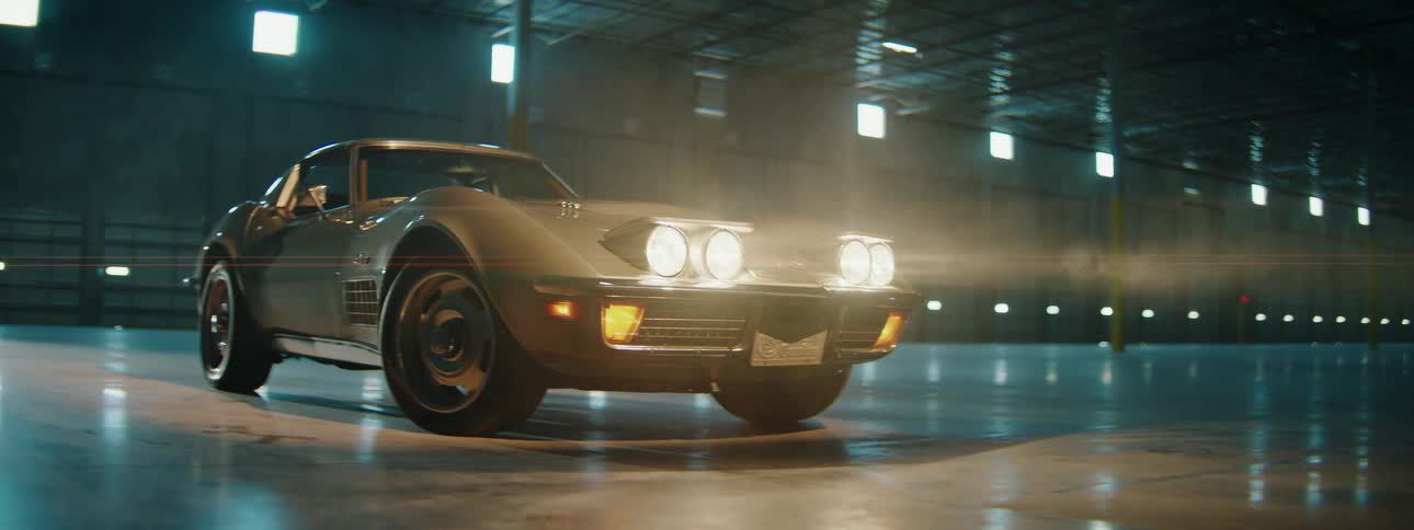 A classic American muscle car is parked in a large, smoky warehouse with its headlights shining brightly