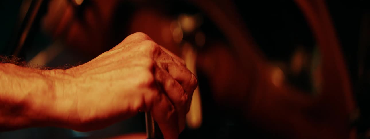 A driver's hand slowly comes to rest and grips a classic car's shiny metal gear shift knob in a dimly lit interior