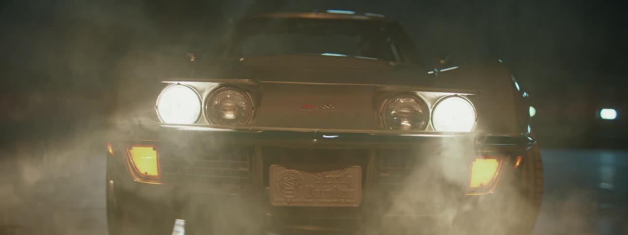 A classic silver Corvette starts up in a smoky garage, with its pop-up headlights flipping open and turning on