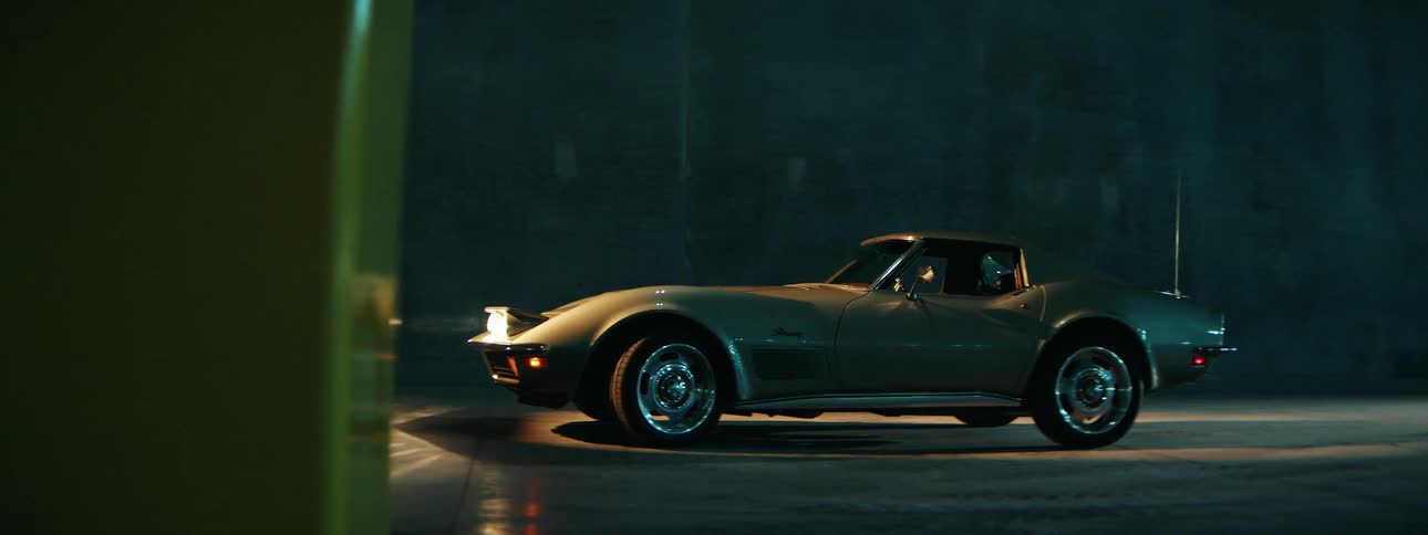 A classic silver sports car with pop-up headlights slowly emerges from the shadows of a dark, moody parking garage at night