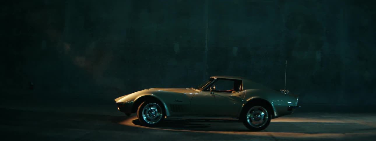 A classic silver Corvette Stingray is parked in a dark, smoky garage under a single spotlight