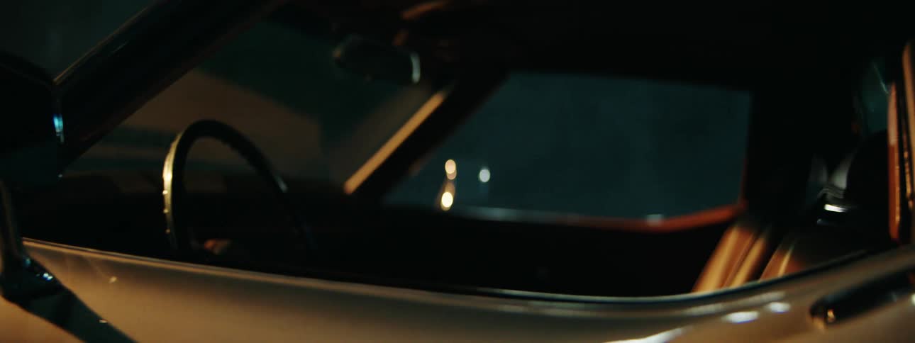 A cinematic close-up shot pans across the sleek body of a vintage silver car parked in a dark, moody environment at night