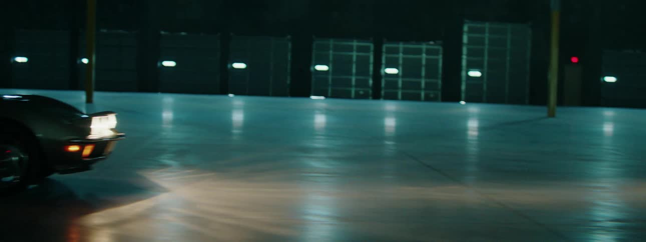 A classic sports car drives through a large, empty, dimly lit warehouse at night