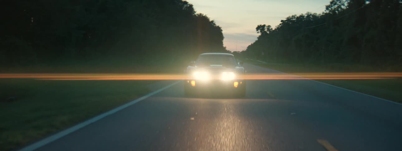 A classic sports car with bright headlights on cruises down a tree-lined country road at dusk with a cinematic lens flare