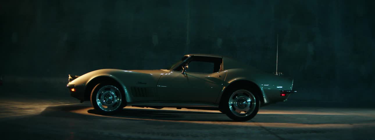 A sleek vintage Corvette Stingray is dramatically lit by a spotlight in a dark, gritty location as it begins to move
