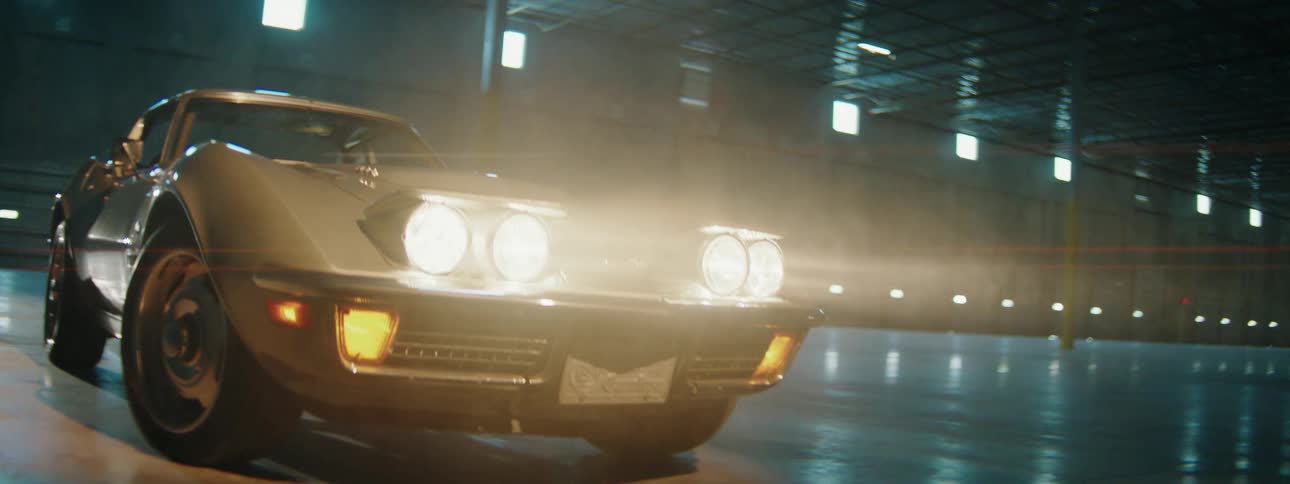 A close-up slow tilt down shot of a classic silver Corvette's pop-up headlights illuminating the smoky air in a large warehouse