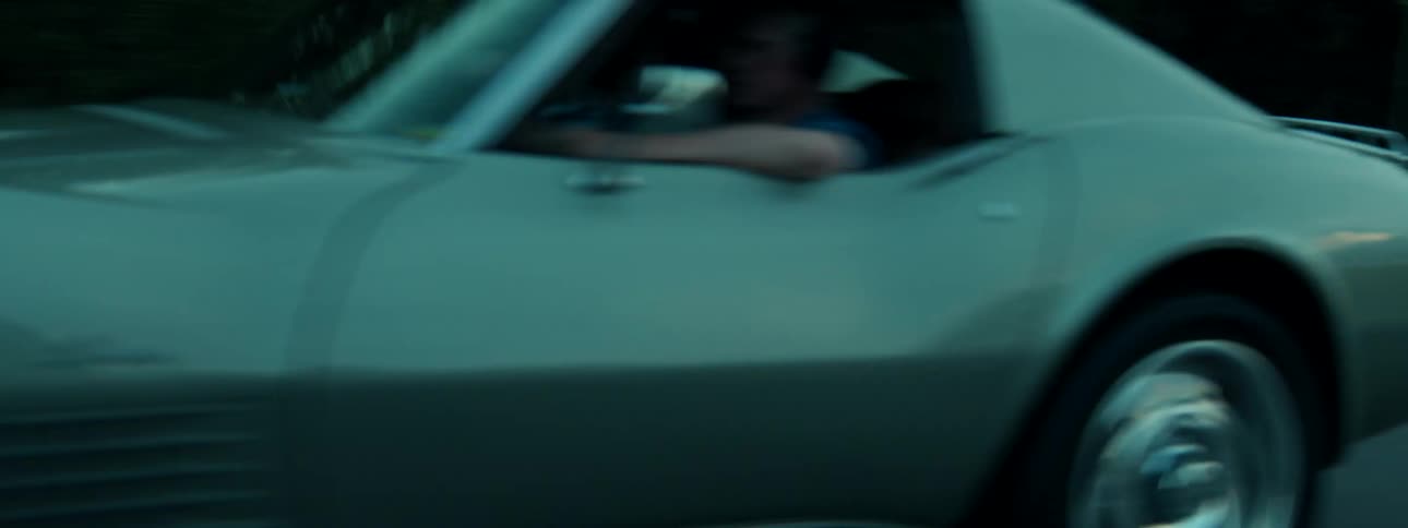 A classic silver sports car with pop-up headlights speeds down a rural highway in the moody, dark light of dusk