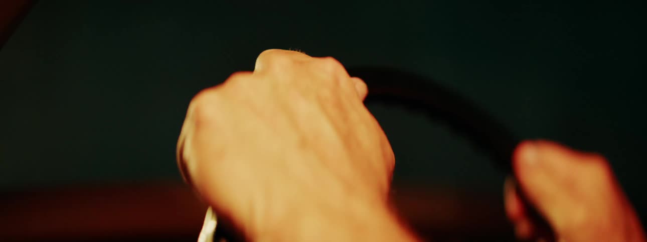 Close-up shot of a driver's hands gripping the steering wheel of a vintage car in a dimly lit, warm environment