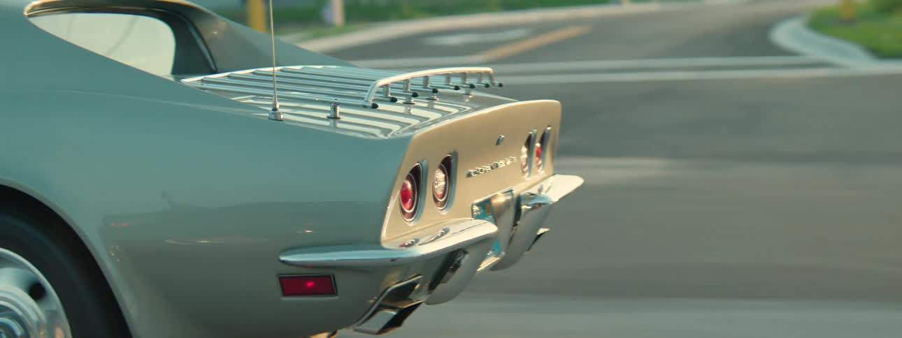A close-up tracking shot follows the rear of a vintage silver Corvette Stingray as it drives along a highway