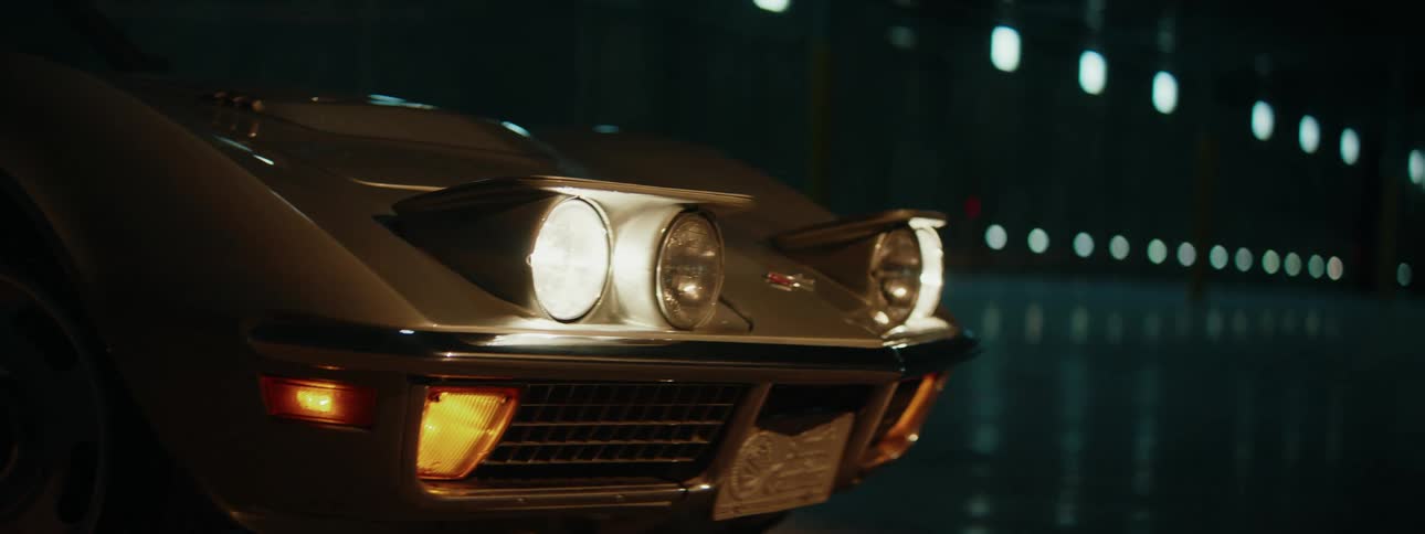 The pop-up headlights of a classic silver Corvette are activated in a dark, atmospheric garage