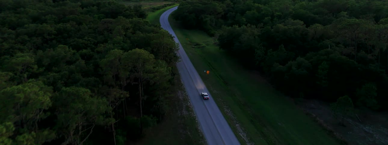 Aerial footage follows a classic white convertible driving along a winding country road through a dense green forest at twilight