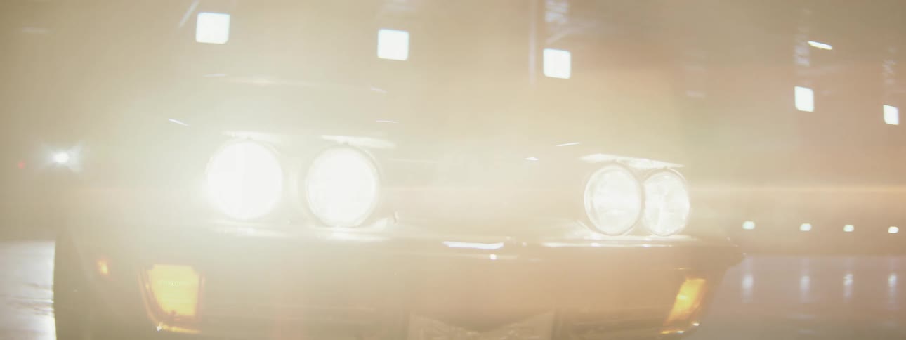 A close-up shot of a classic Corvette's headlights shining brightly through smoke inside a dark warehouse