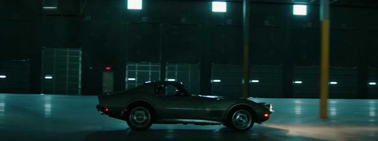 Side profile tracking shot of a vintage Corvette Stingray cruising through a dark, empty industrial warehouse at night