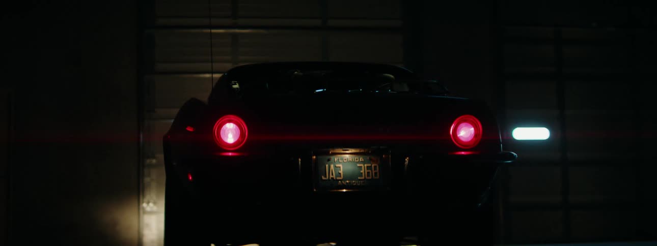A classic black sports car with its red taillights glowing sits parked in a dark garage