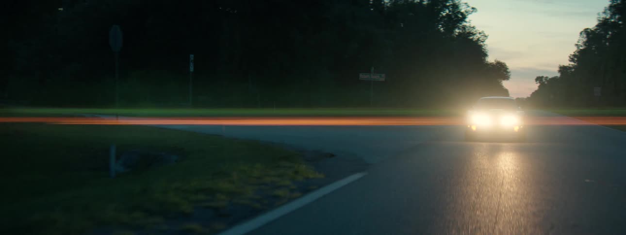 A muscle car with bright headlights sits parked on a rural road at dusk, with a cinematic lens flare stretching across the frame