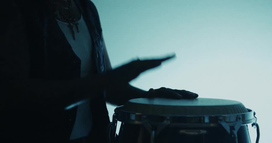 A musician's silhouette is cast as they expertly play a rhythmic beat on a conga drum in a dimly lit room