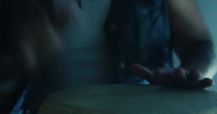 A close-up shot of a musician's hands playing a rhythmic beat on a bongo drum in a dimly lit room