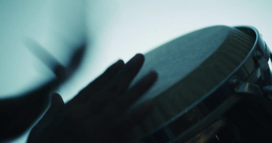 A low angle, silhouetted close-up captures a musician's hands playing a rhythmic beat on a bongo drum