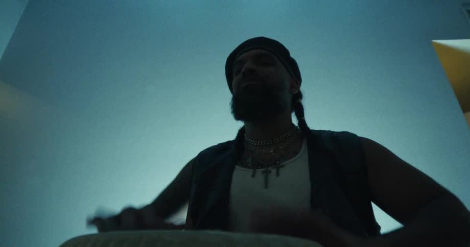 A soulful musician with a beard and beret passionately plays a hand drum, filmed from a creative low-angle perspective in a moody, dimly lit room