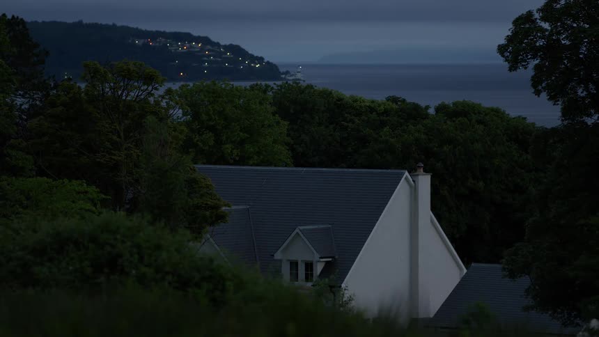 A quiet house nestled in trees overlooks a body of water at dusk as lights from a distant town twinkle on the far shore