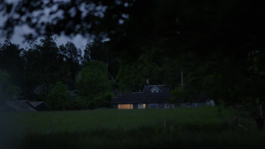 A secluded country house glows with warm light from its windows on a dark evening, viewed from across a field