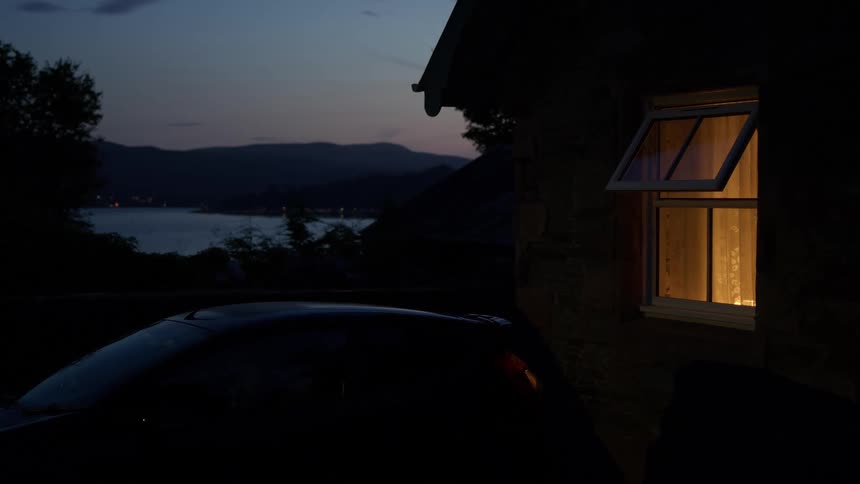 A warmly lit open window on a stone cottage overlooks a tranquil lake at dusk